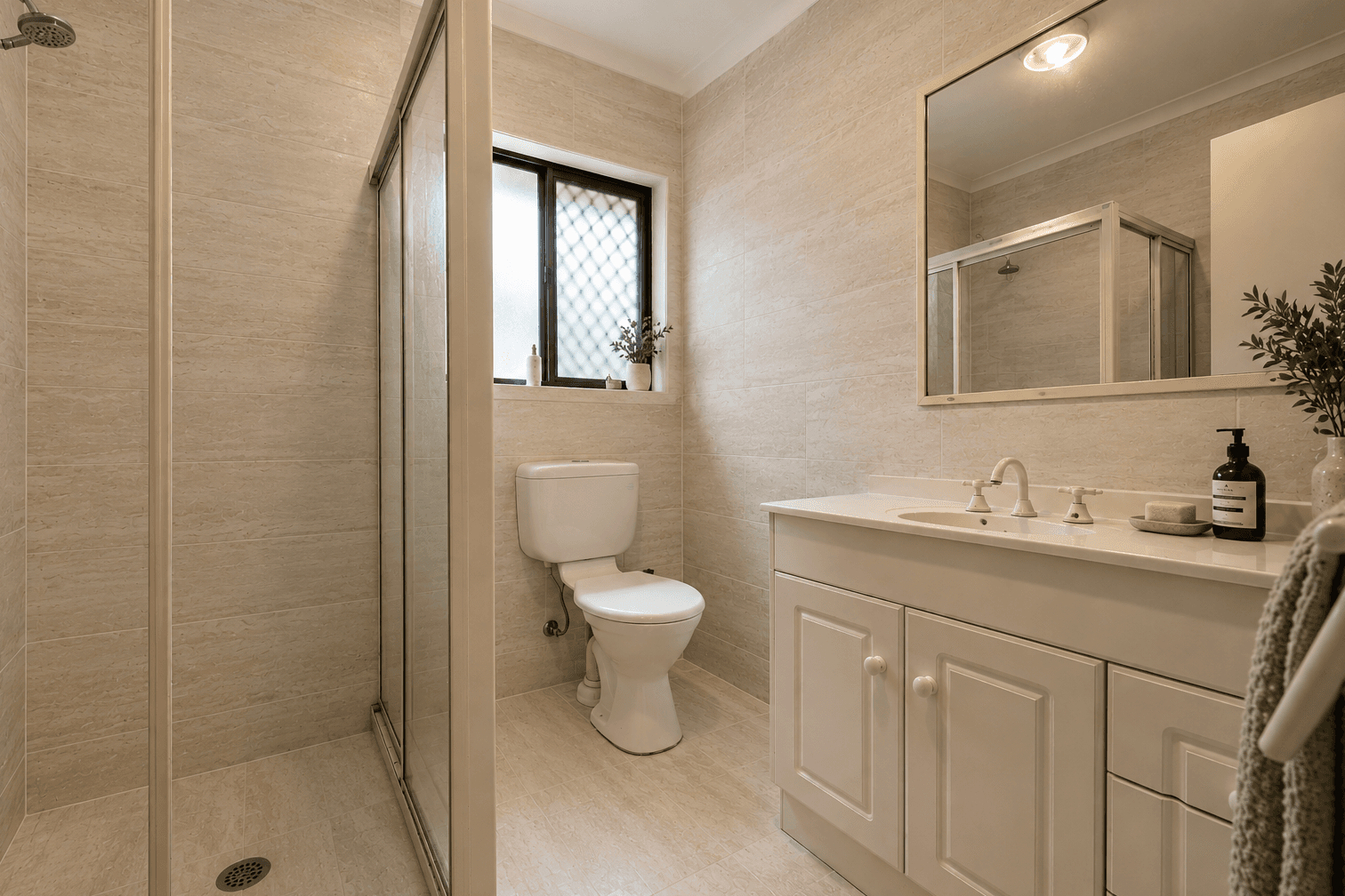 An ensuite, retiled. White tile and floral border out. Warm stone-look walls and floor in. Fittings kept.